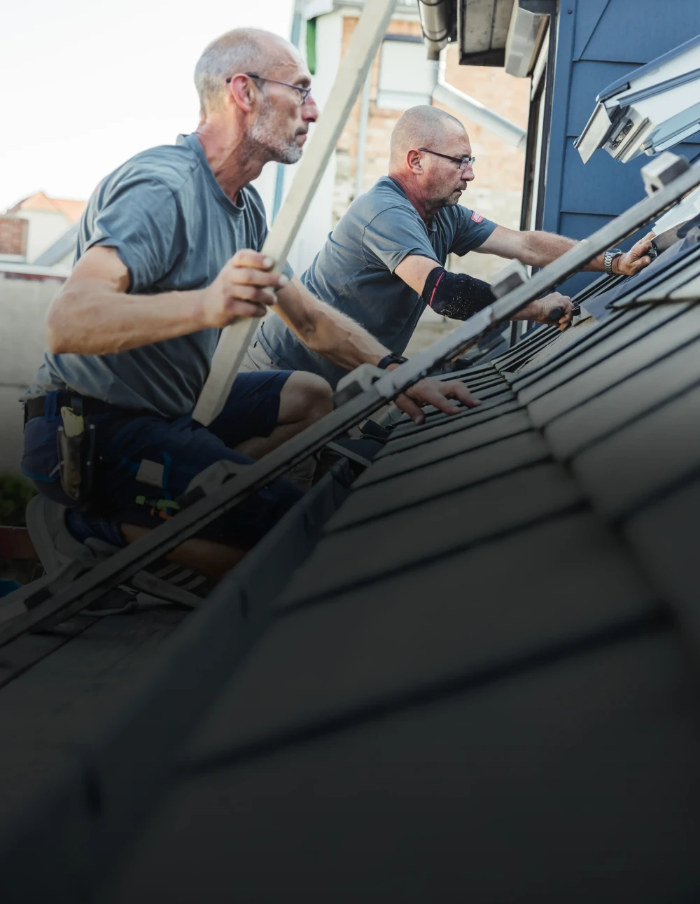Zwei Dachdecker lehnen auf einem Steildach und arbeiten.